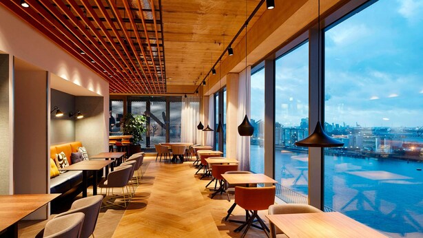 Un área de comedor moderna con mesas y sillas de madera, ventanas amplias con vistas a un paisaje urbano y al agua.