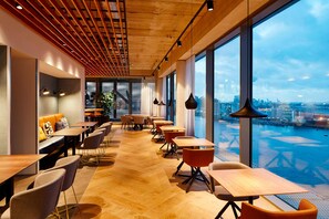 A modern dining area with wooden tables and chairs, large windows with a view of a cityscape and water. - Residence Inn by Marriott Amsterdam Houthavens (Amsterdam)