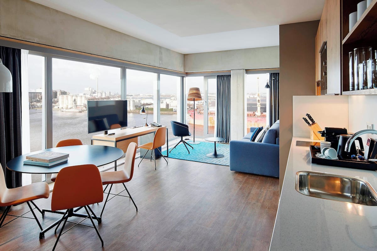 A modern hotel room with a dining area, a sofa, a TV, and a kitchen area with a sink.