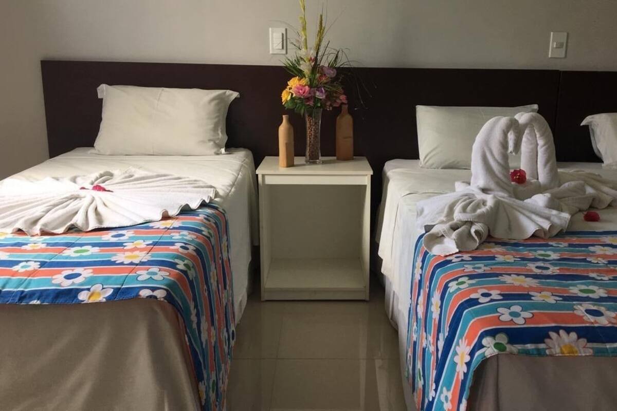 Ein Hotelzimmer mit zwei Betten, jeweils mit floralem Bettbezug und weißen Kissen. Dazwischen befindet sich ein Nachttisch mit Blumen in einer Vase und zwei Flaschen.