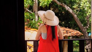 A person wearing a straw hat and a red dress standing by a railing with a view of trees and a tiled roof. - Pousada Murici (Barreirinhas)