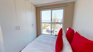 A bedroom with a large window, white wardrobe, and red pillows. - Mont Blu Condo (Cape Town)
