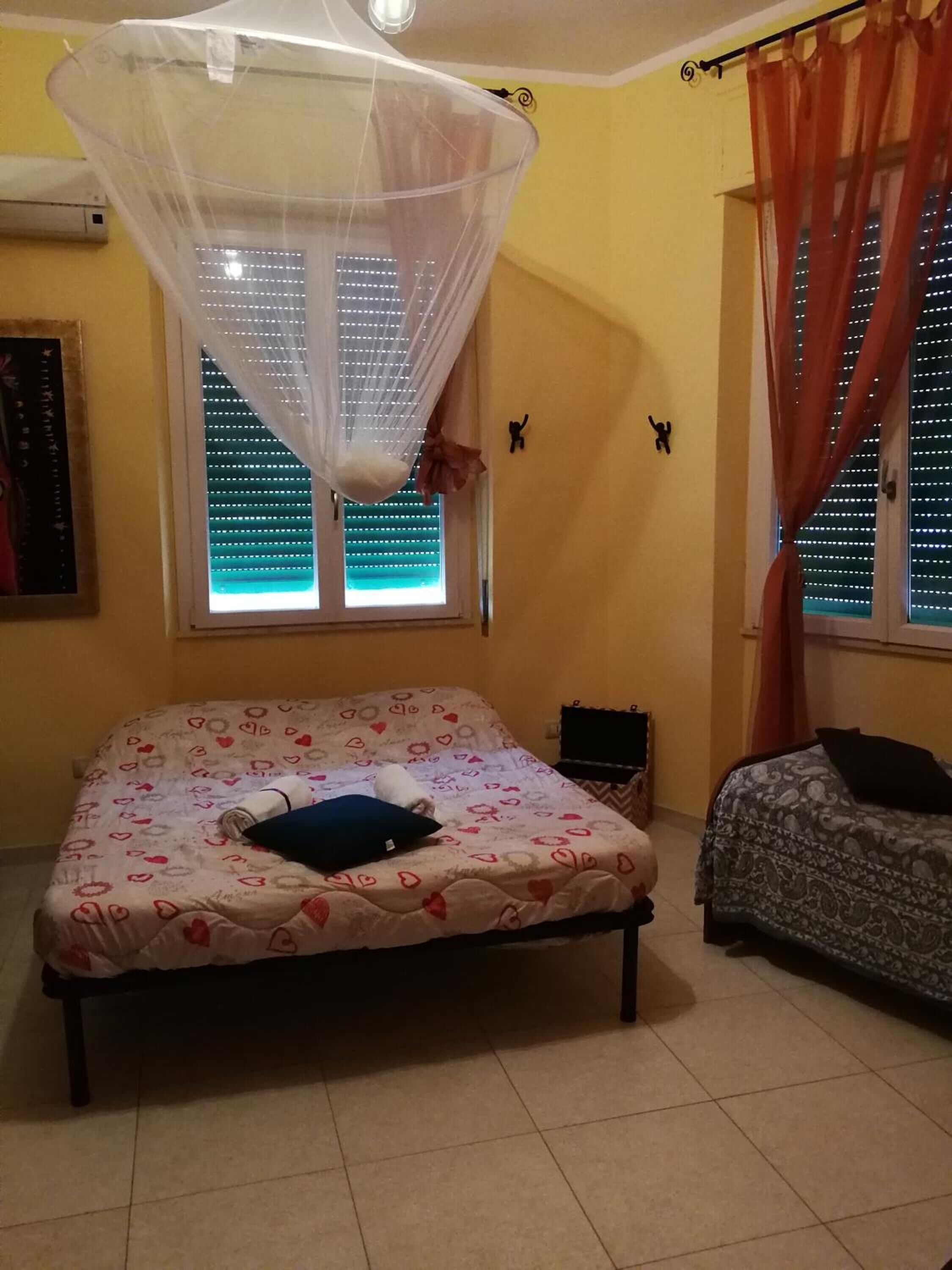 A bedroom with a bed, a bedside table, a window with blinds, and a mosquito net.