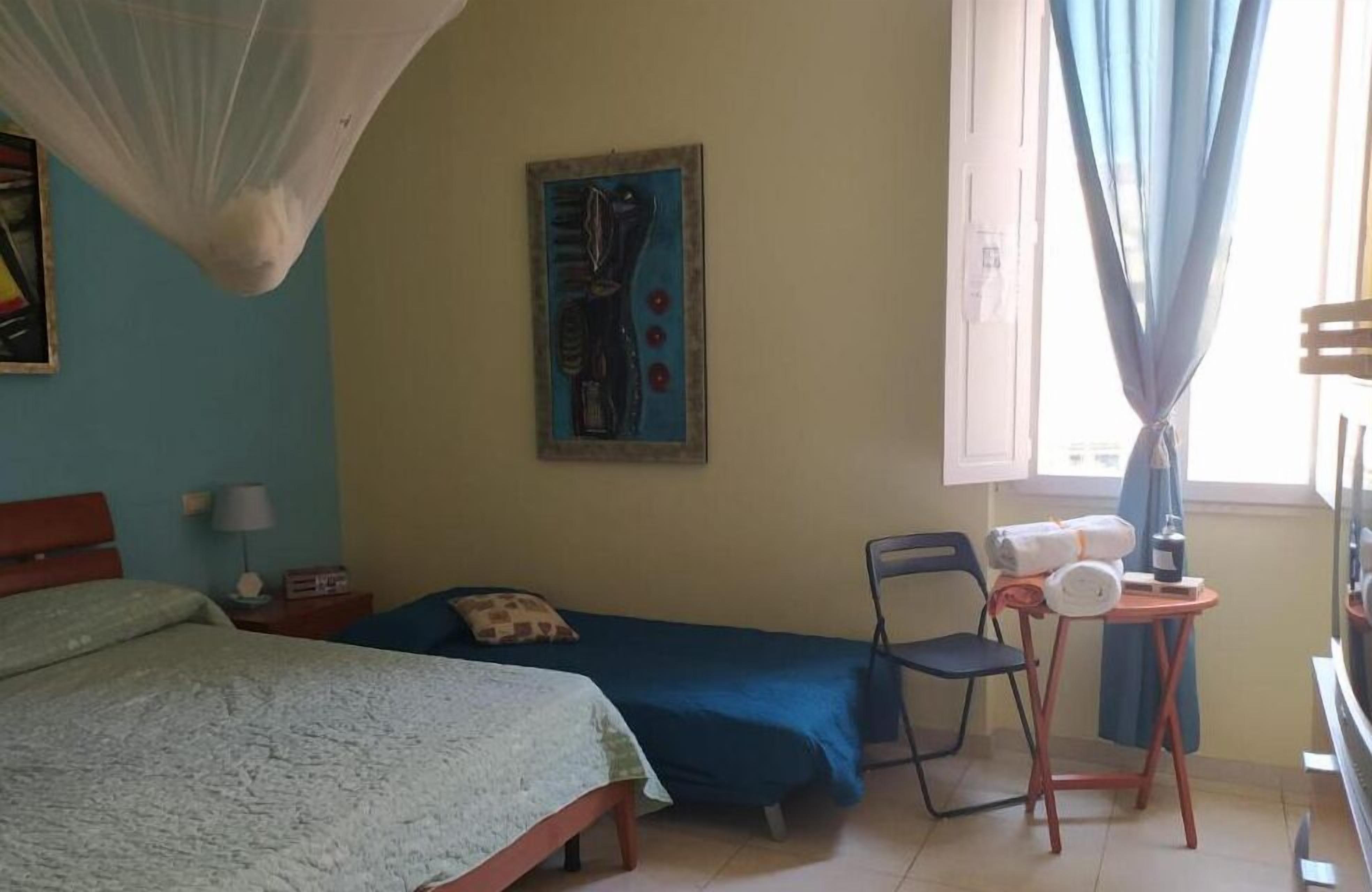 A room with two single beds, a chair, a small table with items, a painting on the wall, and a window with curtains.