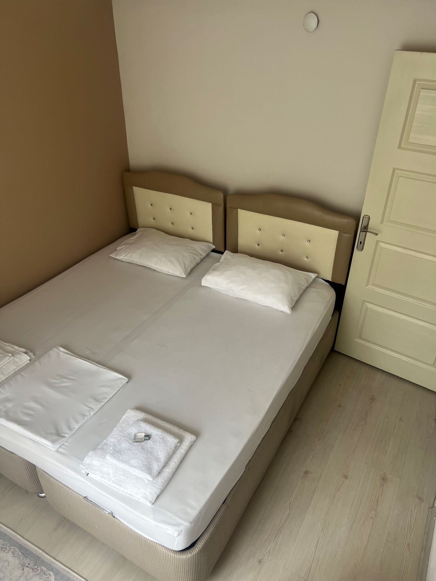 A double bed with a beige headboard and white bedding in a room with light wooden flooring and a closed door.