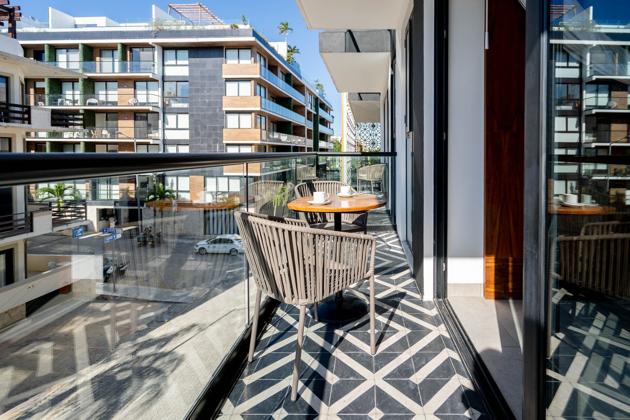 Un appartement moderne doté d’un balcon, d’un coin repas avec chaises et table, et offrant une vue sur d’autres immeubles et une rue.