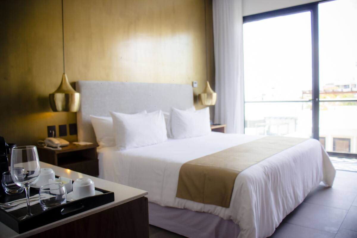 Habitación de hotel con una cama grande, una mesita de noche con lámpara, una bandeja con vasos y una taza, y vista al exterior a través de un amplio ventanal.
