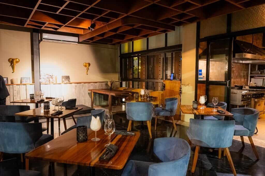 A restaurant with wooden tables and blue chairs, a kitchen visible through the glass wall, and decorative wall art.
