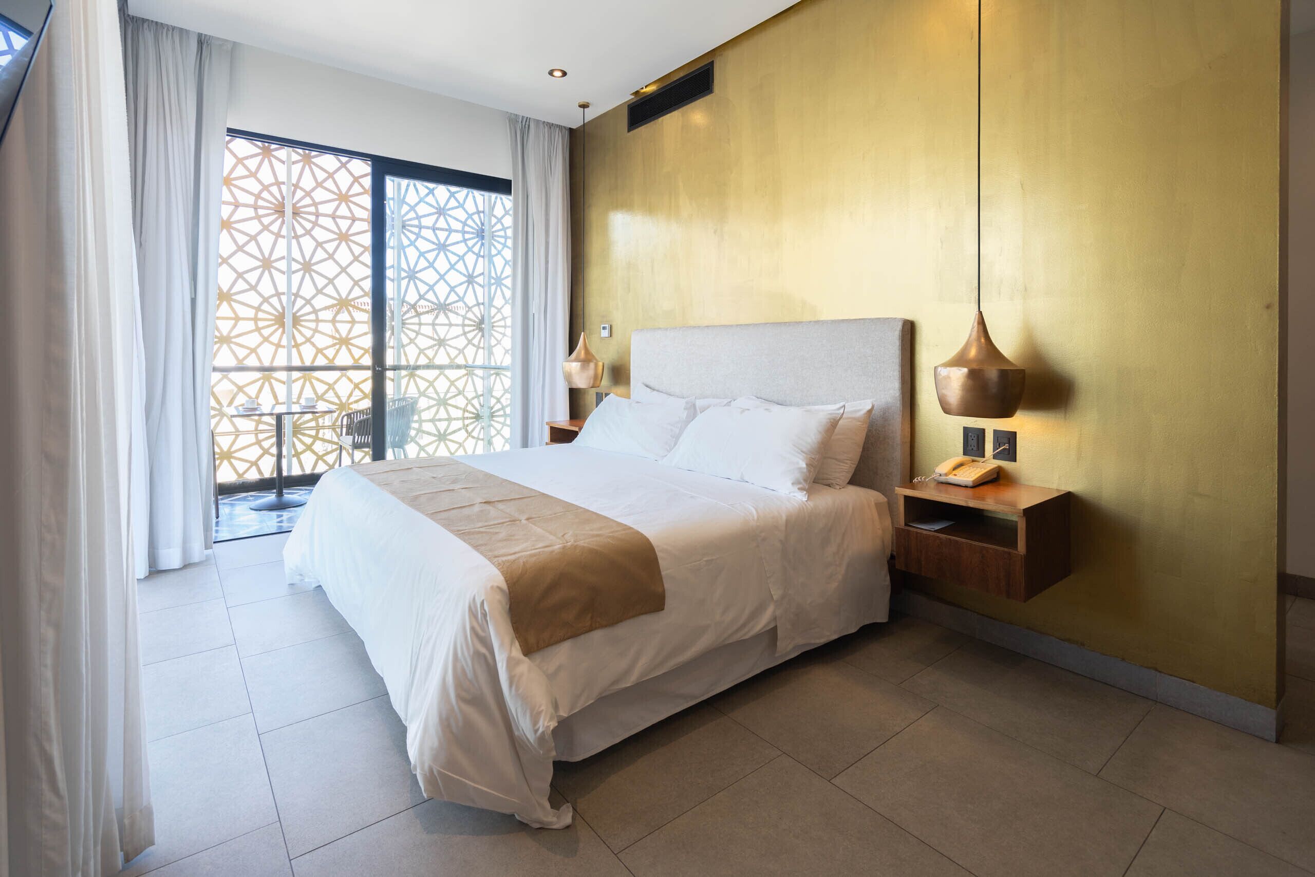 A hotel room with a large bed, a nightstand, a lamp, and a view of a balcony with a geometric patterned door.