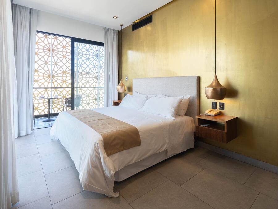 Une chambre d’hôtel avec un grand lit, une table de chevet, une lampe et une vue sur un balcon dont la porte est ornée d’un motif géométrique.