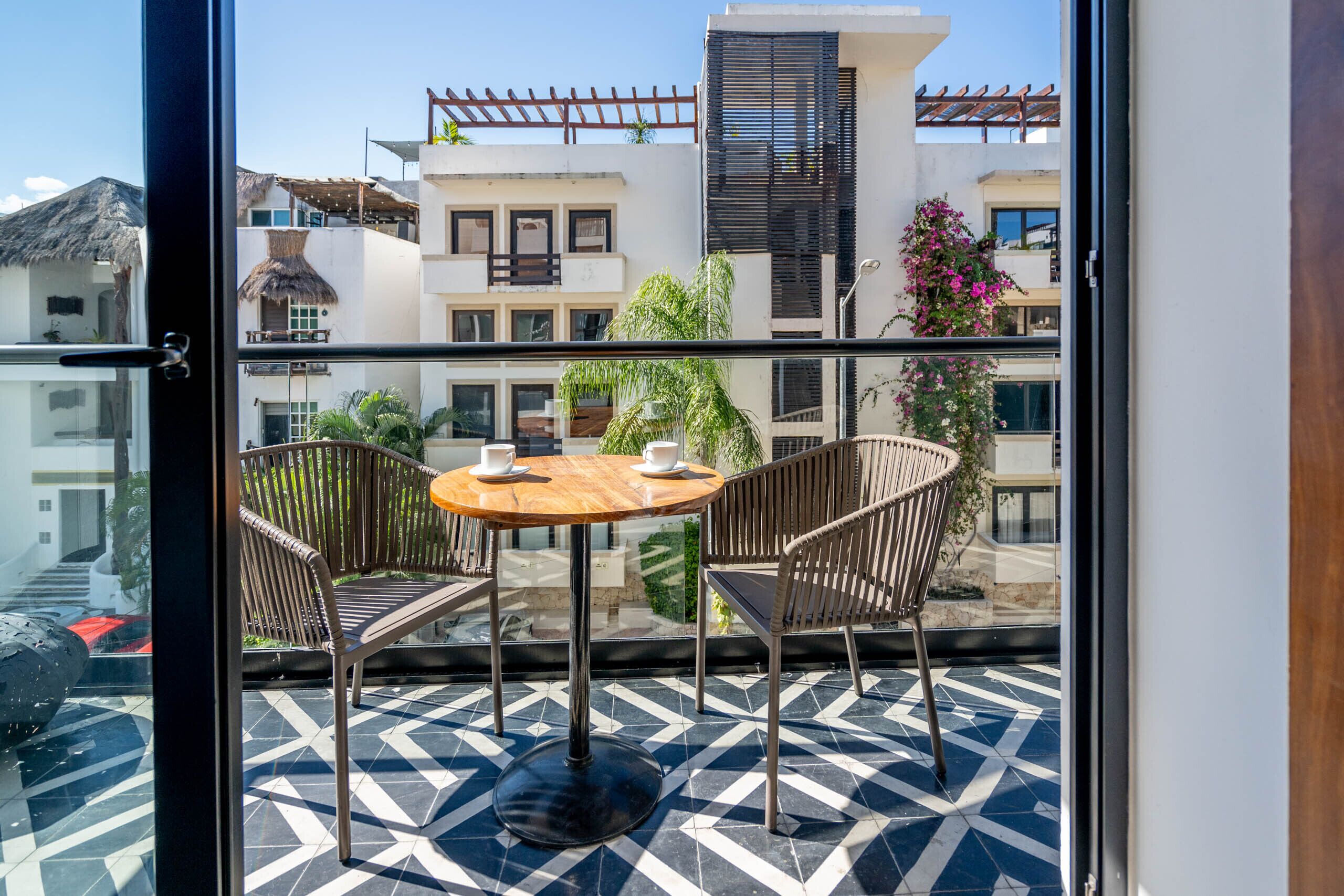 Un balcon avec une table ronde et deux chaises, un sol à motifs et une vue sur des immeubles dotés de balcons et de plantes.