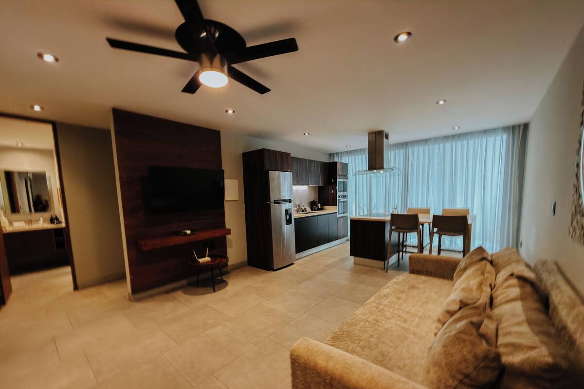 Una sala de estar moderna con un sofá, un televisor, un ventilador de techo y una zona de cocina con un refrigerador y una mesa de comedor.