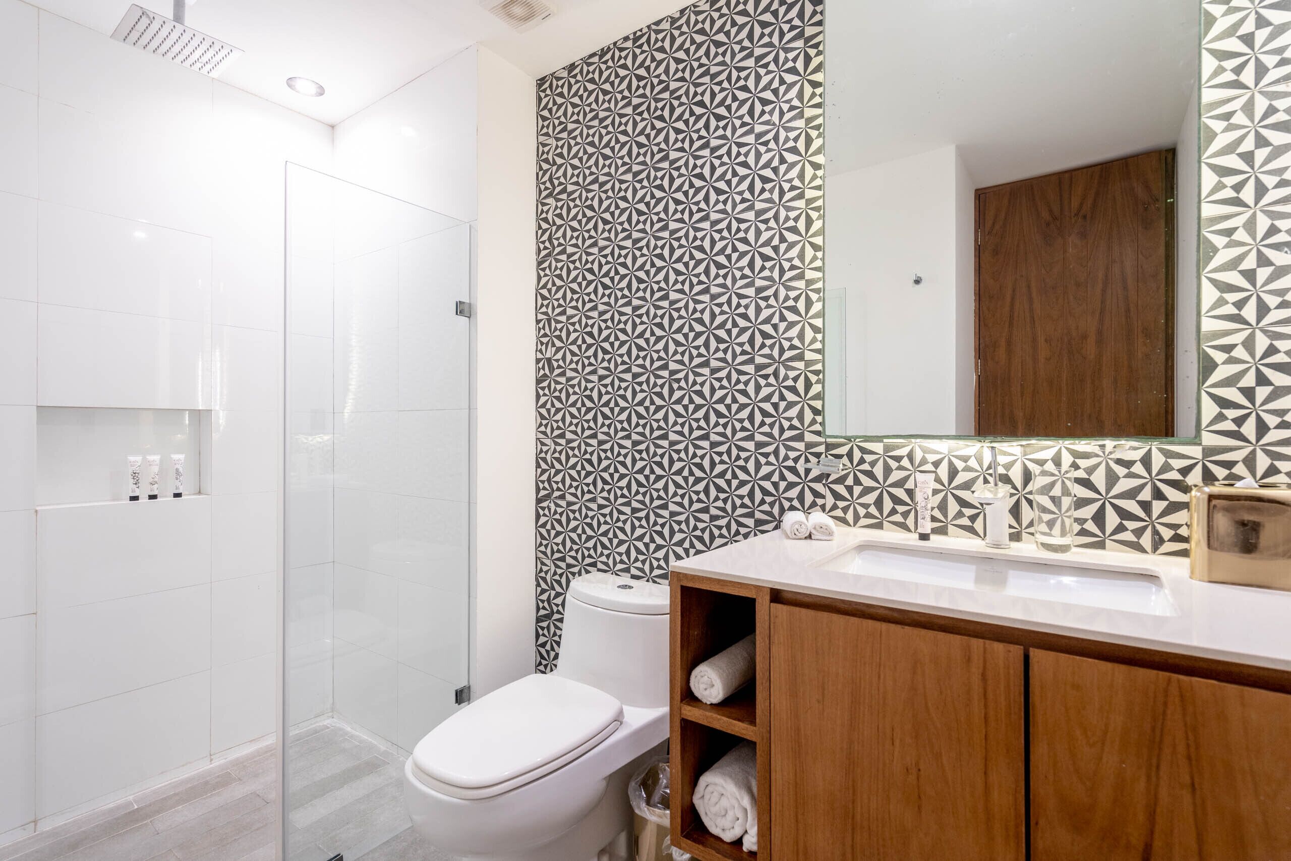 A modern bathroom with a white toilet, a shower enclosure, a wooden vanity with a mirror, and patterned tiles on the walls.