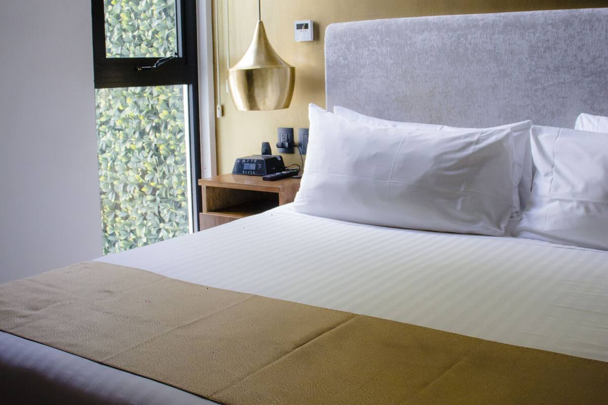 Habitación de hotel con una cama grande, una mesita de noche con lámpara y ventana con vistas a zonas verdes.