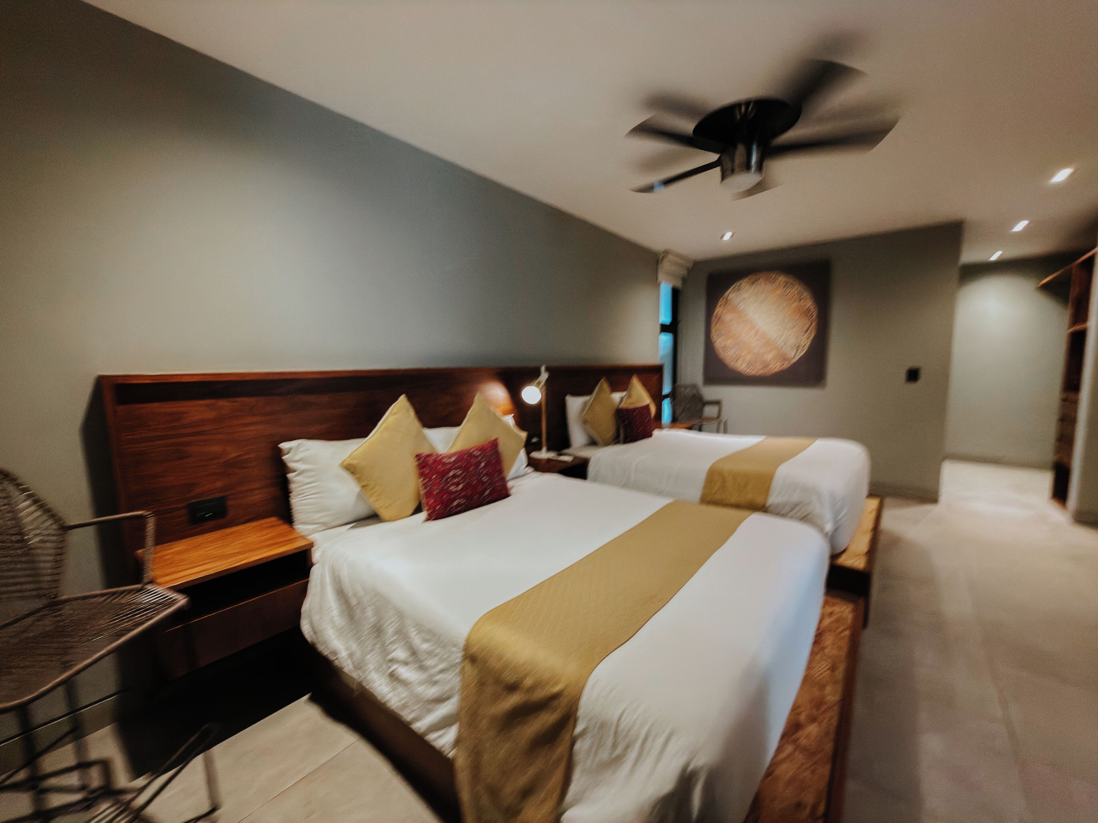 A hotel room with two beds, a ceiling fan, a nightstand, a chair, and a wall-mounted lamp.