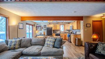 A spacious living room with a large sofa, a coffee table with a puzzle, and a dining area with wooden cabinets and a kitchen in the background.