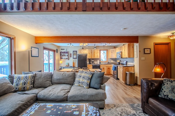 A spacious living room with a large sofa, a coffee table with a puzzle, and a dining area with wooden cabinets and a kitchen in the background. - Private Cottage Nestled in the Trees;Pet Friendly-Near Slopes-Waterpks-JimThorpe (Albrightsville)