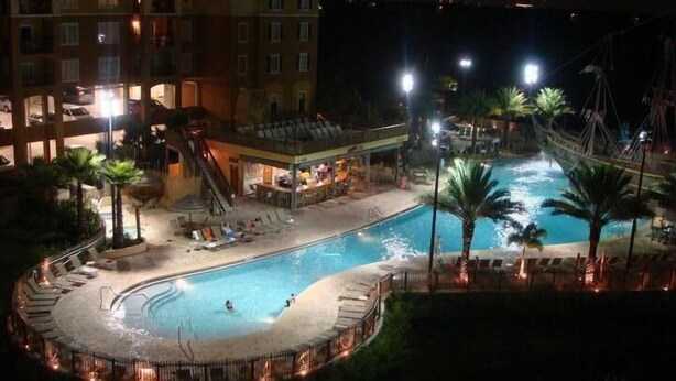 Vista nocturna de un área de piscina de hotel con una fuente circular, sillas de descanso y palmeras.