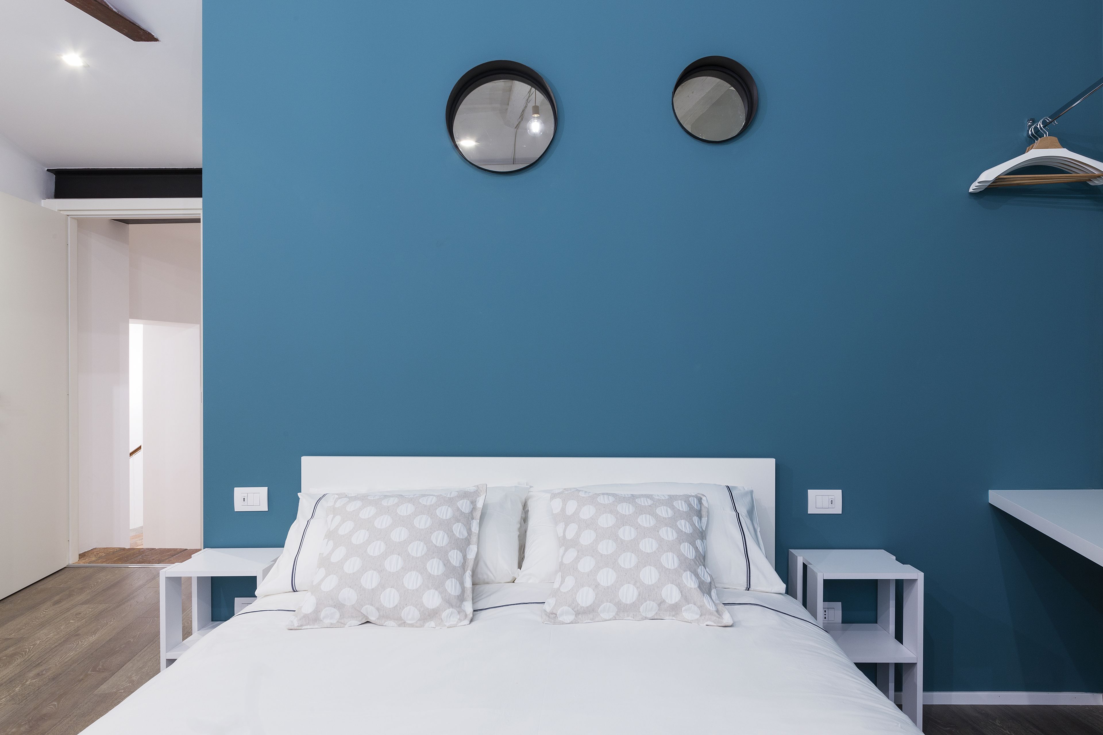 A modern bedroom with a blue accent wall, a bed with white bedding and patterned pillows, two round mirrors on the wall, and a white nightstand.