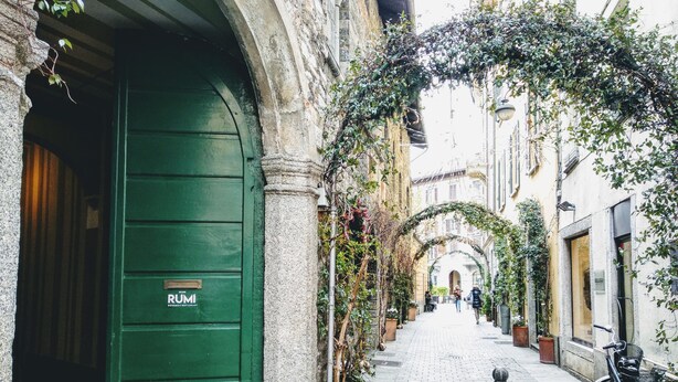 A narrow cobblestone street with a green door labeled "RUMI" on the left, flanked by buildings with ivy-covered arches and potted plants.