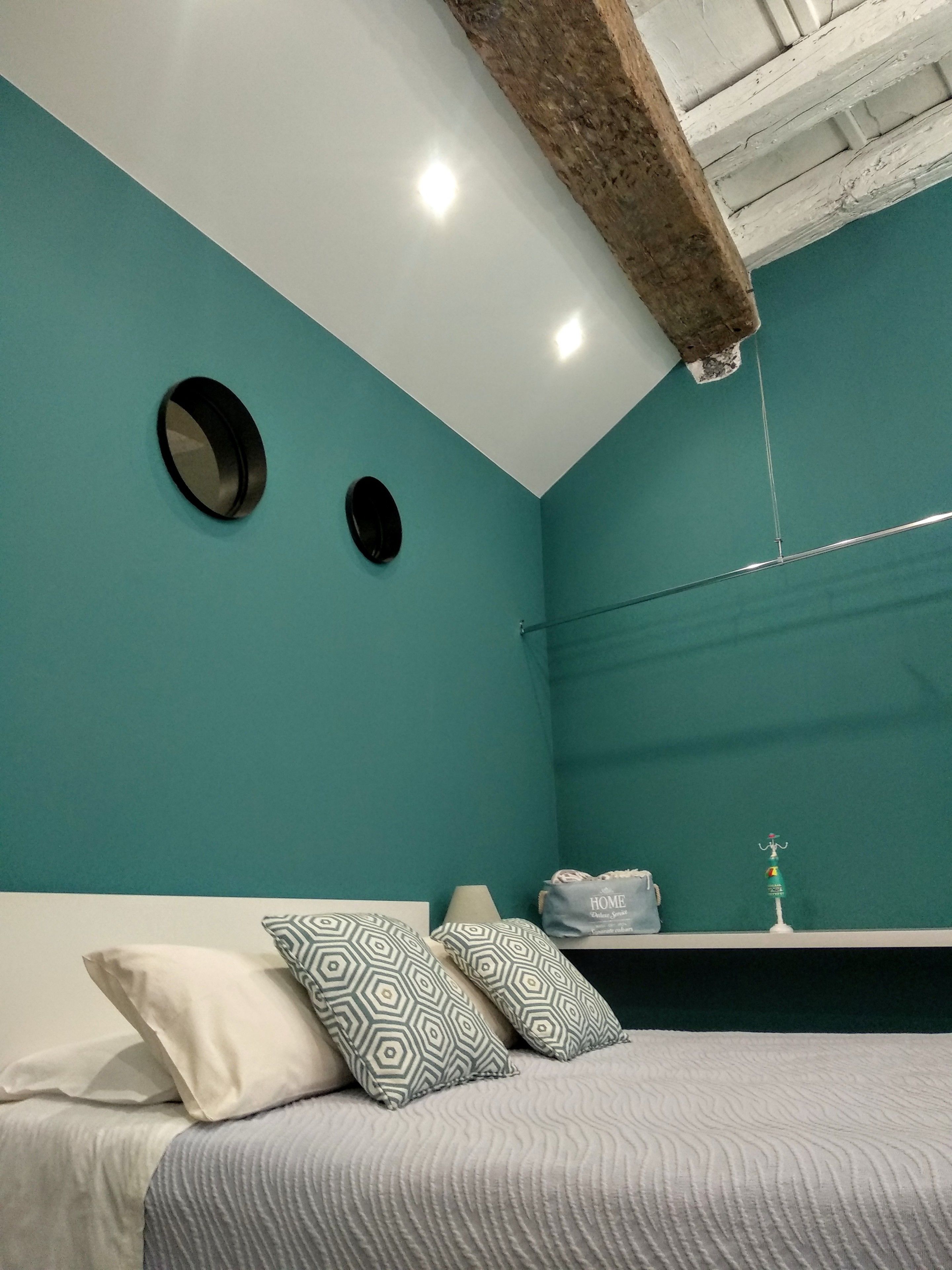 A bedroom with teal walls, a bed with patterned pillows, a round mirror, and a hanging light fixture.