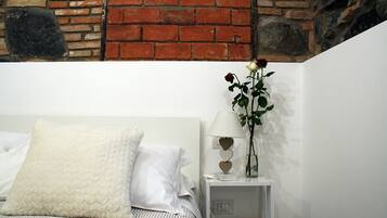 Ein ordentlich bezogenes Bett mit weißen Leinen und einem großen weißen Kissen, ein Nachttisch mit Lampe und einer Vase mit einer einzelnen Rose, sowie eine Backsteinwand im Hintergrund.