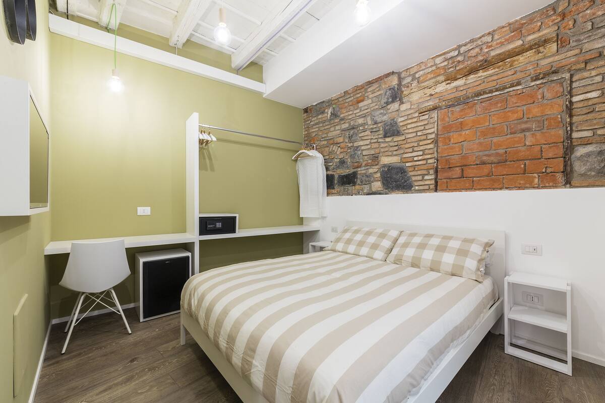 Un dormitorio con cama, escritorio, silla, un pequeño armario y una pared de ladrillo.
