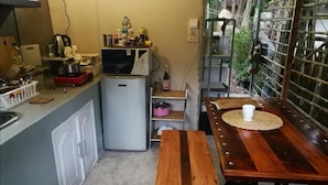 A compact kitchen with a microwave, refrigerator, and various kitchen items on the counter. There is a wooden bench with a woven placemat and a white mug on it. - Brookhouse at Khaoyai (Pak Chong)