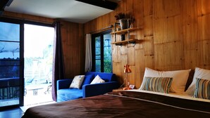 A room with a bed, a blue sofa, and a wooden wall. There is a window with curtains, a shelf with items, and a view outside. - Brookhouse at Khaoyai (Pak Chong)