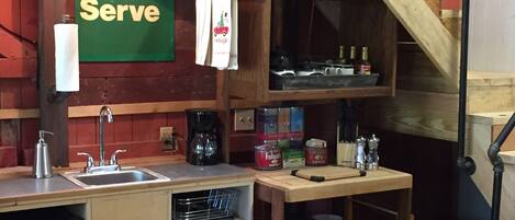 Une petite cuisine équipée d’un évier, d’une machine à café et d’étagères de rangement. On peut voir une pancarte indiquant « Service complet », et une pompe à essence Texaco est fixée au mur.