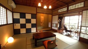A traditional Japanese room with tatami flooring, a low table with cushions, sliding doors, and a view through large windows.