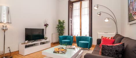 A modern living room with a flat-screen TV, teal armchairs, a brown sofa, and a white coffee table.