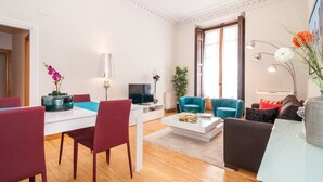 A modern living room with a white sofa, a blue armchair, and a dining table with red chairs. There is a TV, a lamp, and a vase with flowers on the table. - MADRID-SOL-GATE PLAZA MAYOR, New Apartment, Comodo, modern design. (Madrid)