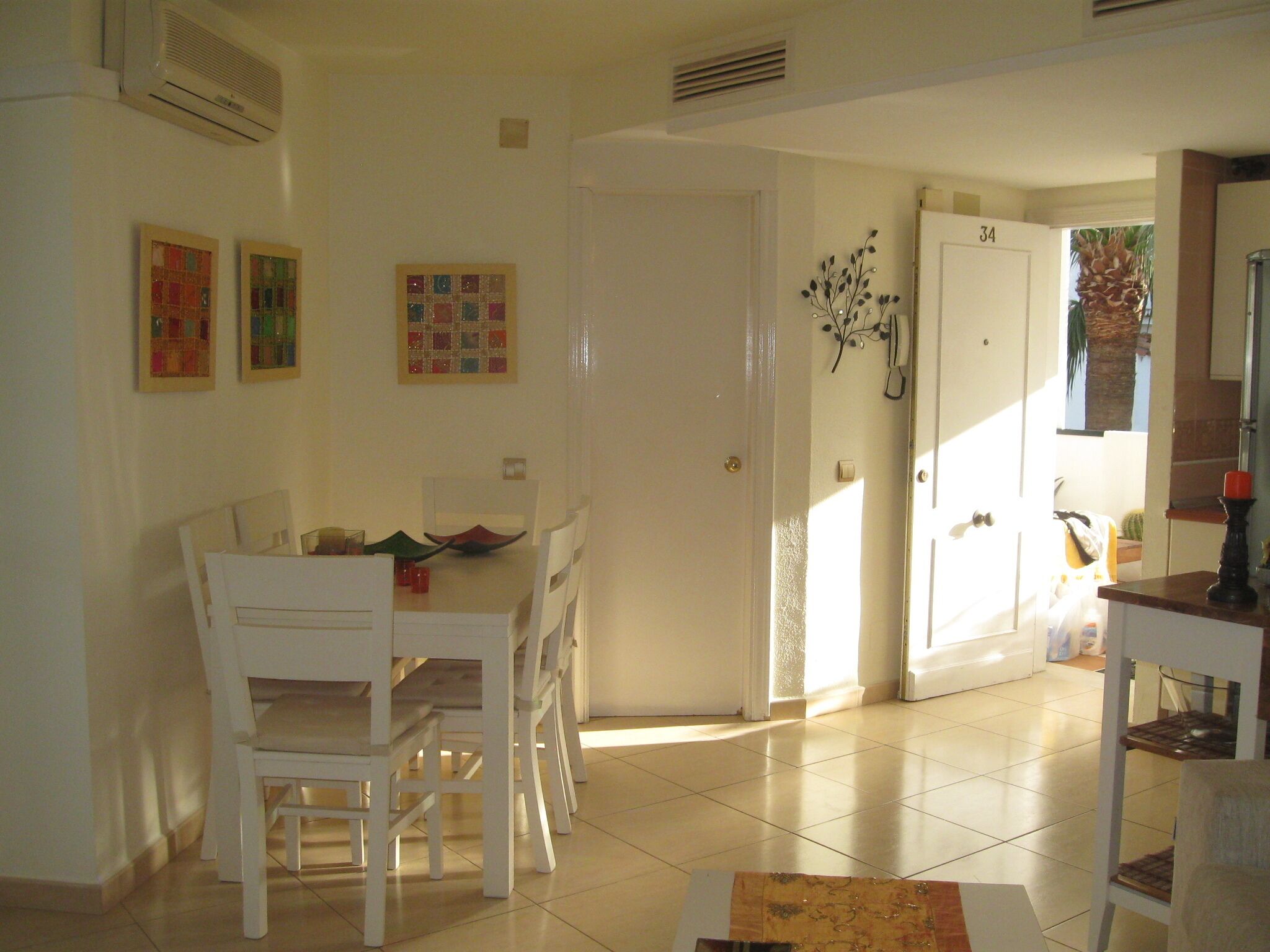 Un comedor con una mesa y sillas blancas, piso de baldosas y una puerta que da a otra habitación.