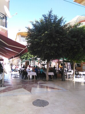 Un área de comedor al aire libre con mesas y sillas, un árbol y un edificio con un letrero.