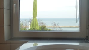 A bathroom with a bathtub, a window with a view of the sea, and a towel hanging on the window.