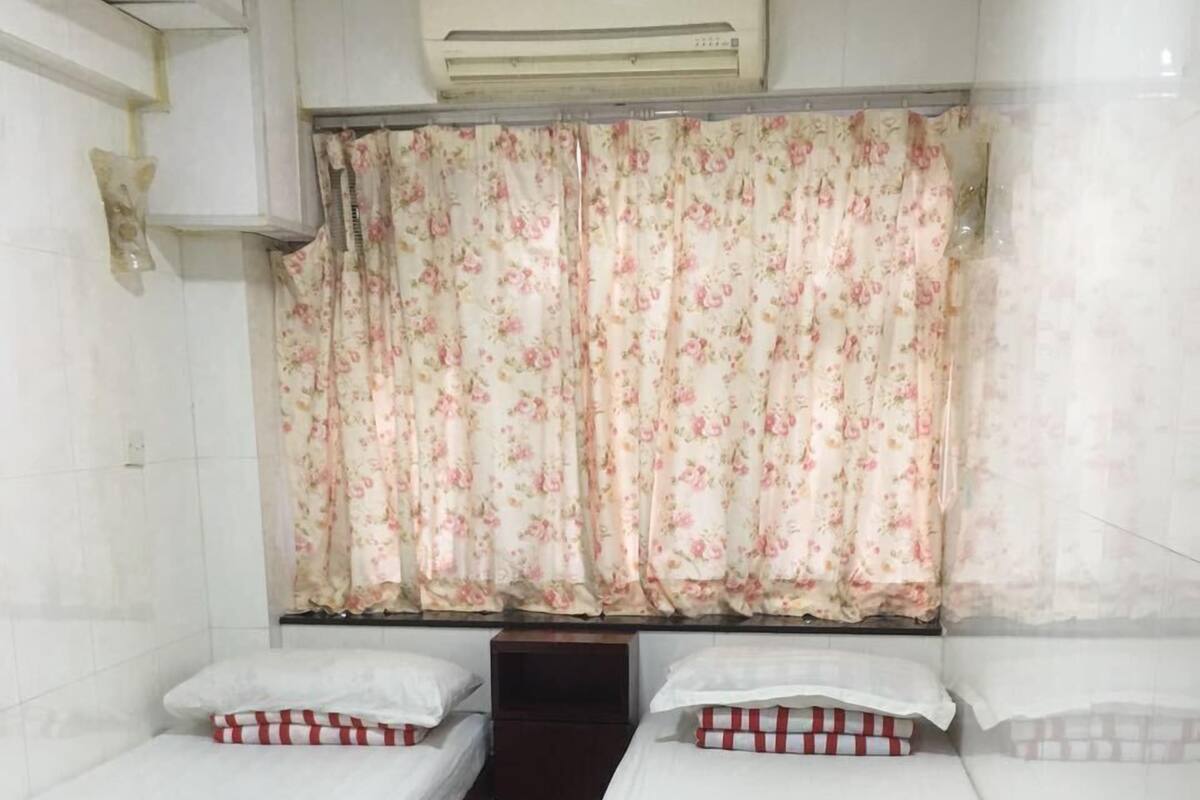 A room with two single beds, each with a wooden frame and white bedding. There is a small wooden nightstand between the beds, and a wall-mounted air conditioning unit above.