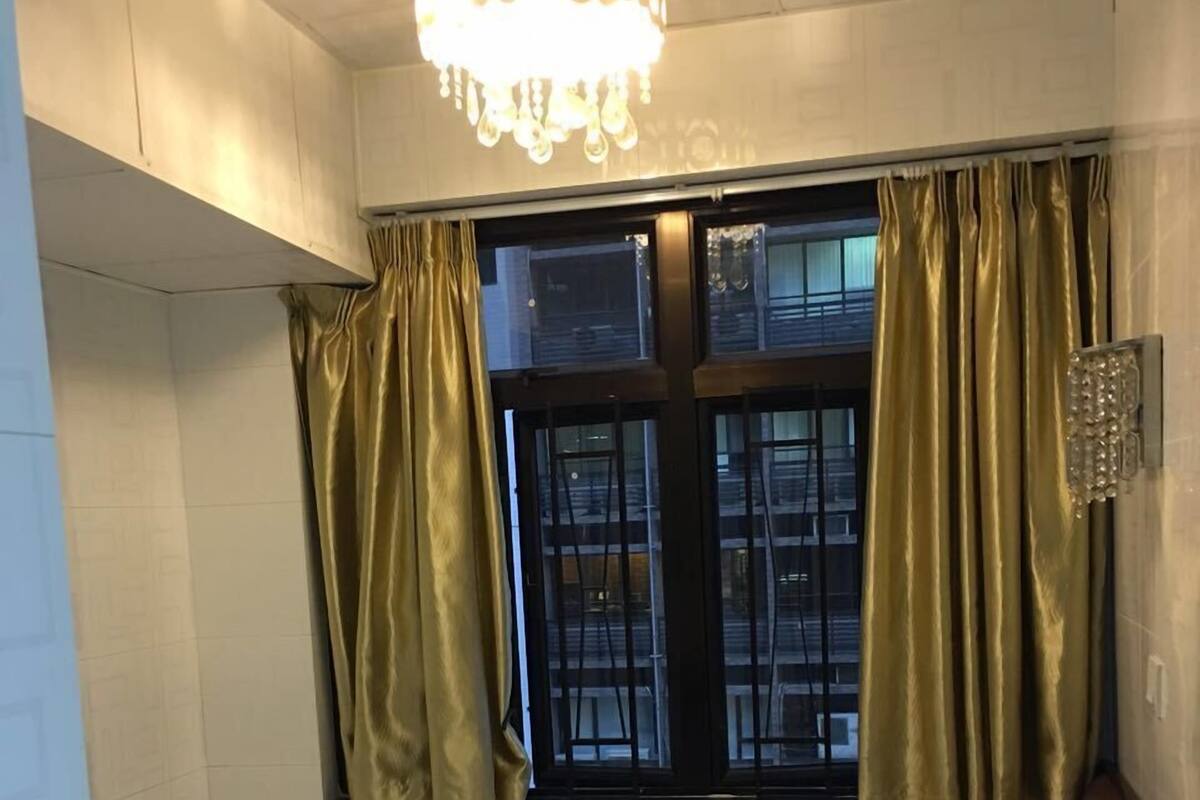A small, single-bed room with a window, curtains, and a chandelier.