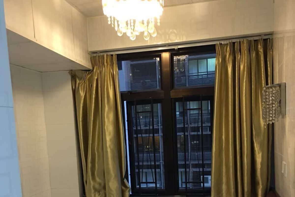 A small, single-bed room with a window, curtains, and a chandelier.