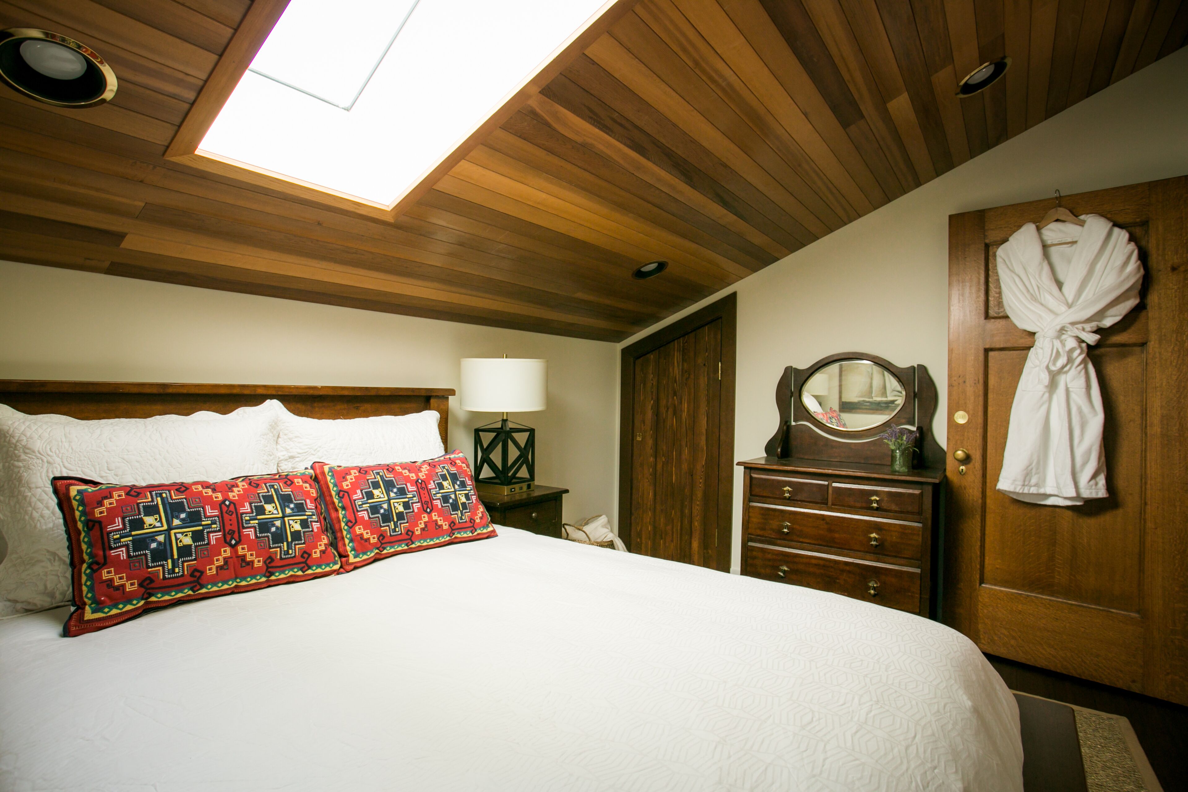 Un dormitorio con techo de madera, una cama con ropa de cama blanca, un cojín estampado en rojo y negro, un tocador de madera, un espejo y una bata colgada en la puerta.