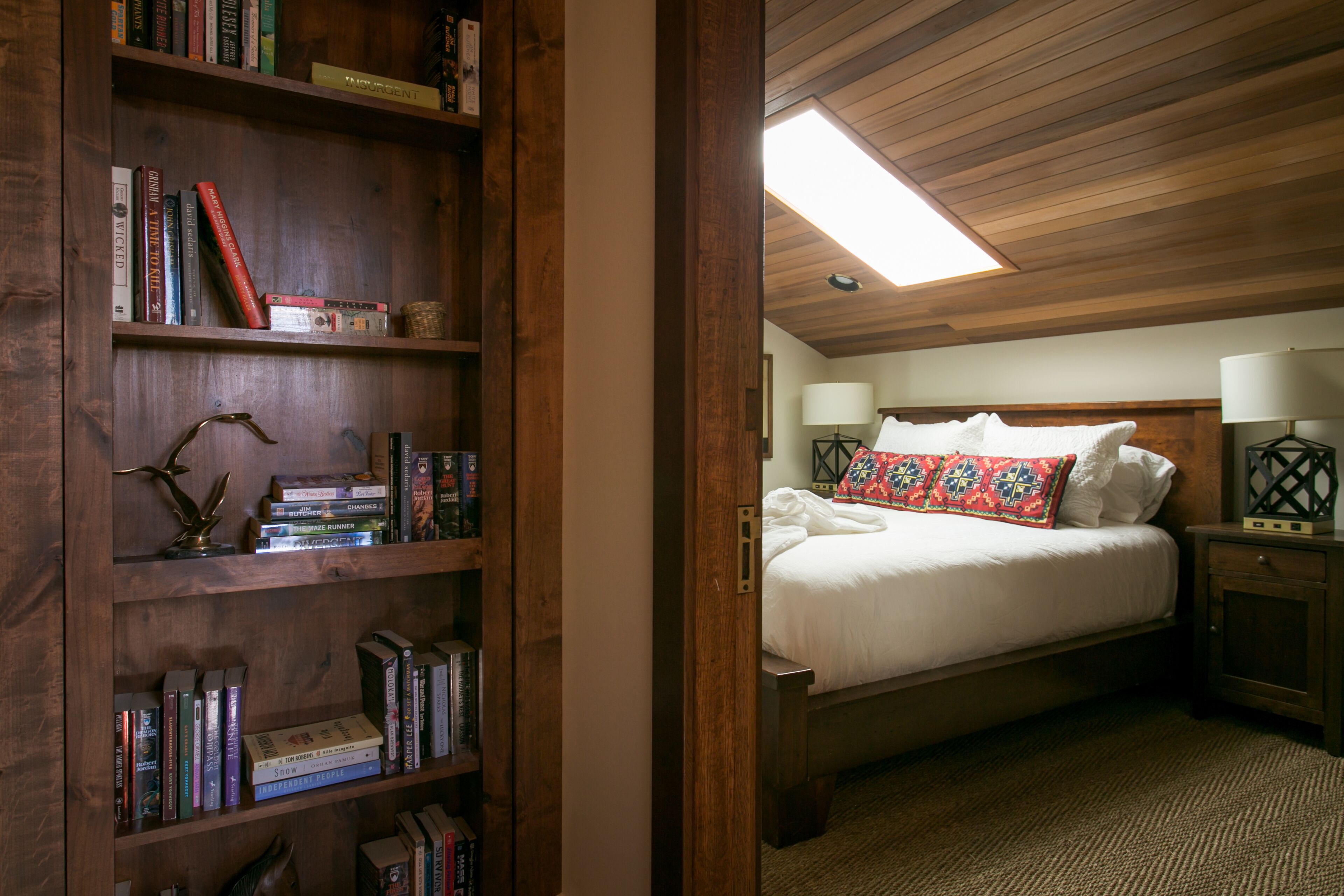 Une chambre avec une bibliothèque en bois, un lit recouvert d’une literie blanche et orné d’un coussin coloré, une table de chevet avec une lampe, et une lucarne au-dessus.