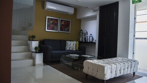 Una sala de estar moderna con un sofá, un reposapiés y una mesa de centro de vidrio. Se ve una escalera con una planta en maceta, y una obra de arte en la pared.