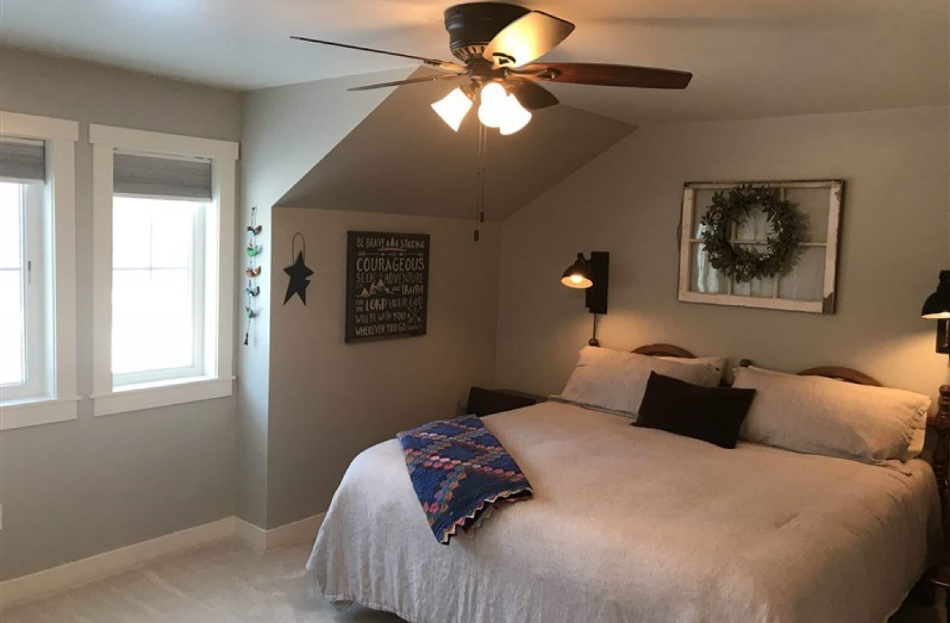 Un dormitorio con cama, ventilador de techo, lámpara de pared, ventana y un adorno en la pared.