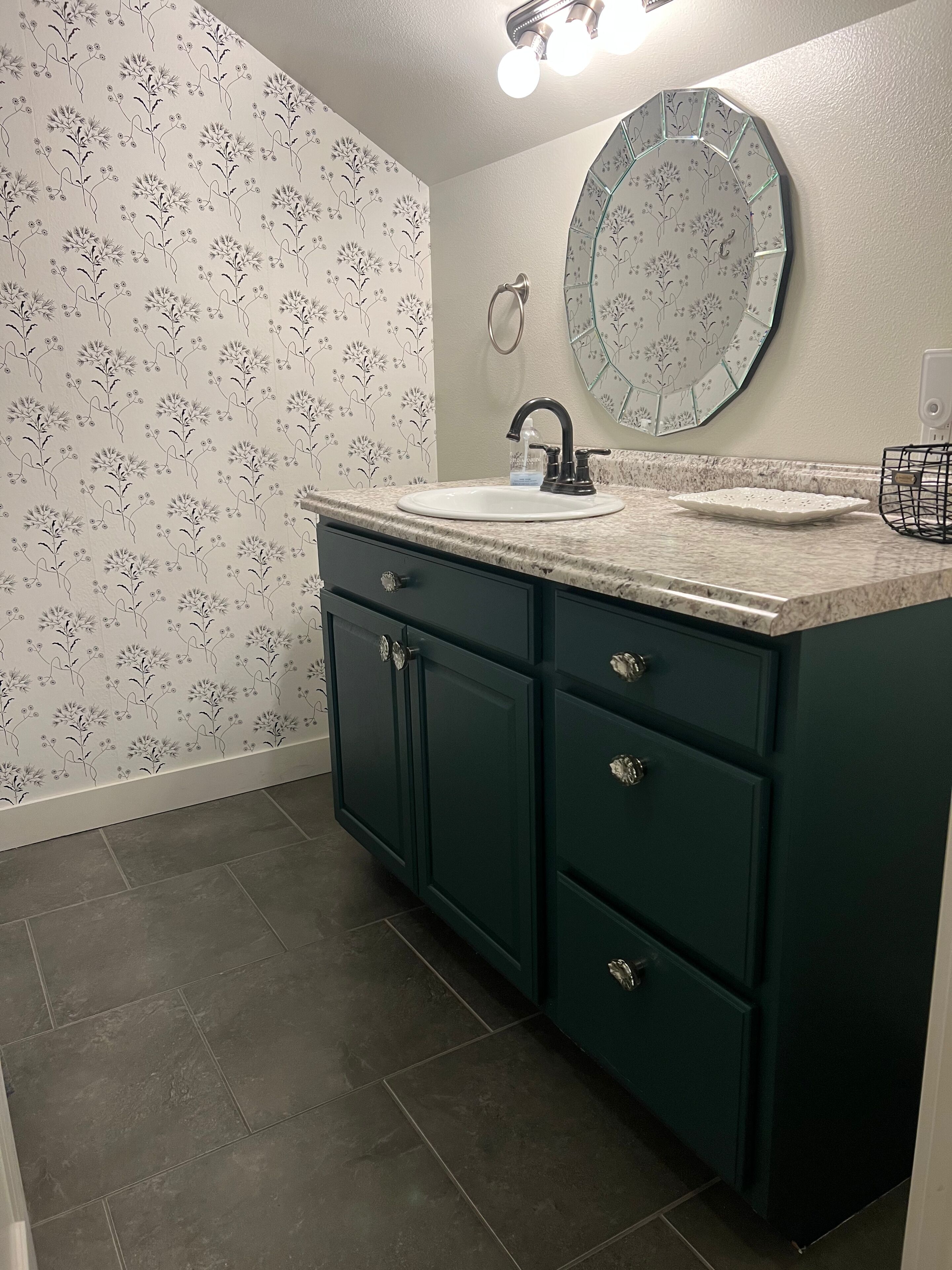 A bathroom with a sink, dark green cabinets, and a floral wallpaper pattern.