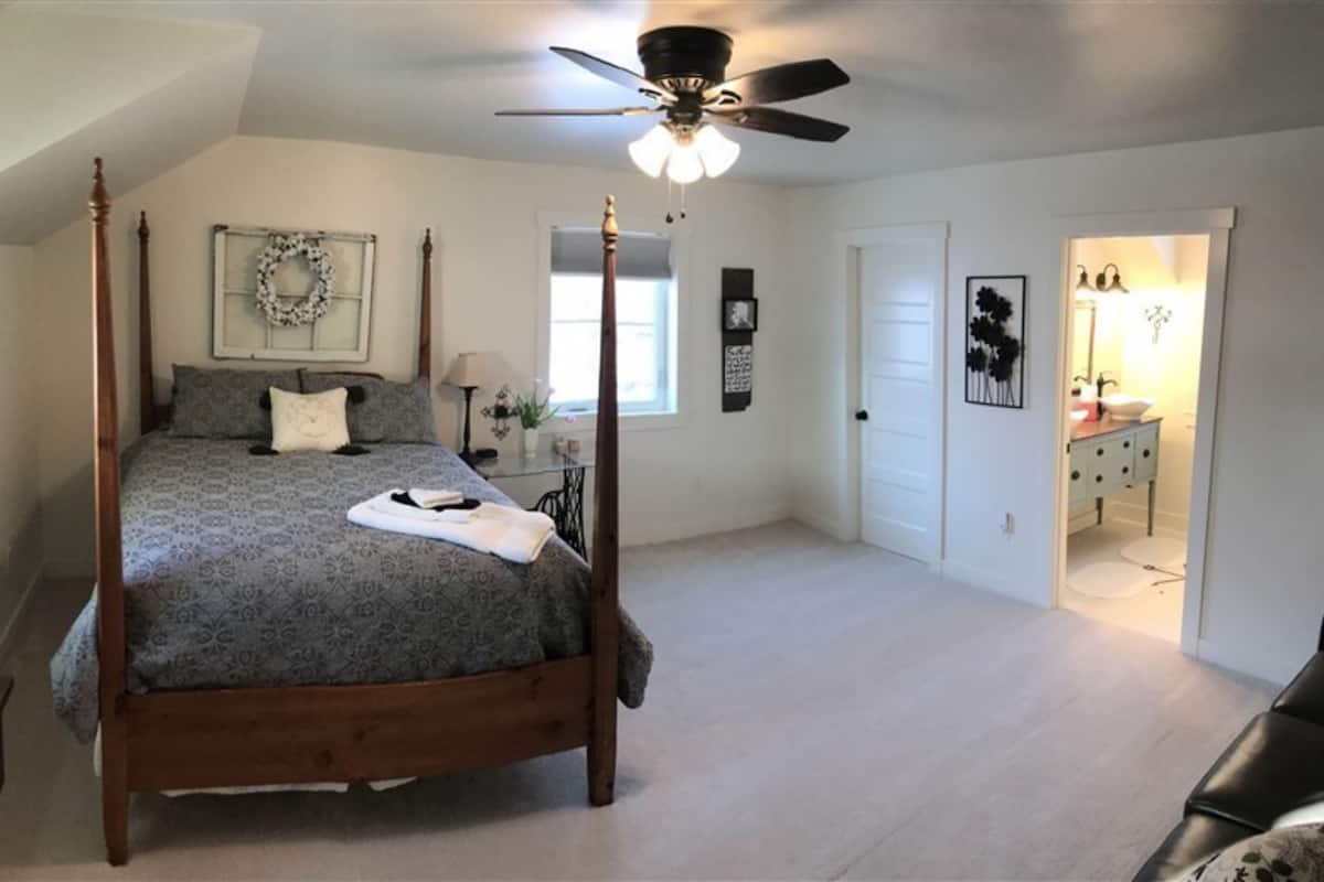 A four-poster bed in a bedroom with a ceiling fan, a window with a wreath, and a door leading to a bathroom.