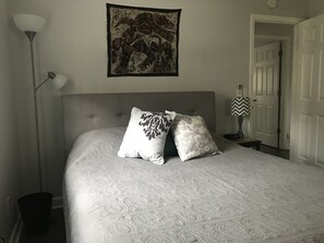 A bedroom with a bed, two pillows, a nightstand with a lamp, a framed picture on the wall, and a door in the background. - Cute Cottage, Pet and Family Friendly, Reduced Rates for  Longer Term Rentals (Black Mountain)