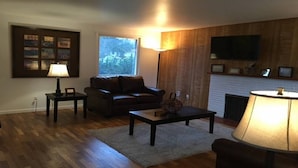 Un salón con sofá, mesa de centro y lámpara. Hay una chimenea y un televisor montado en la pared.