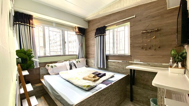 A bedroom with a bed, a bedside table, a window with curtains, a plant, and a wooden wall. - Hong Kong Hostel (Kowloon)