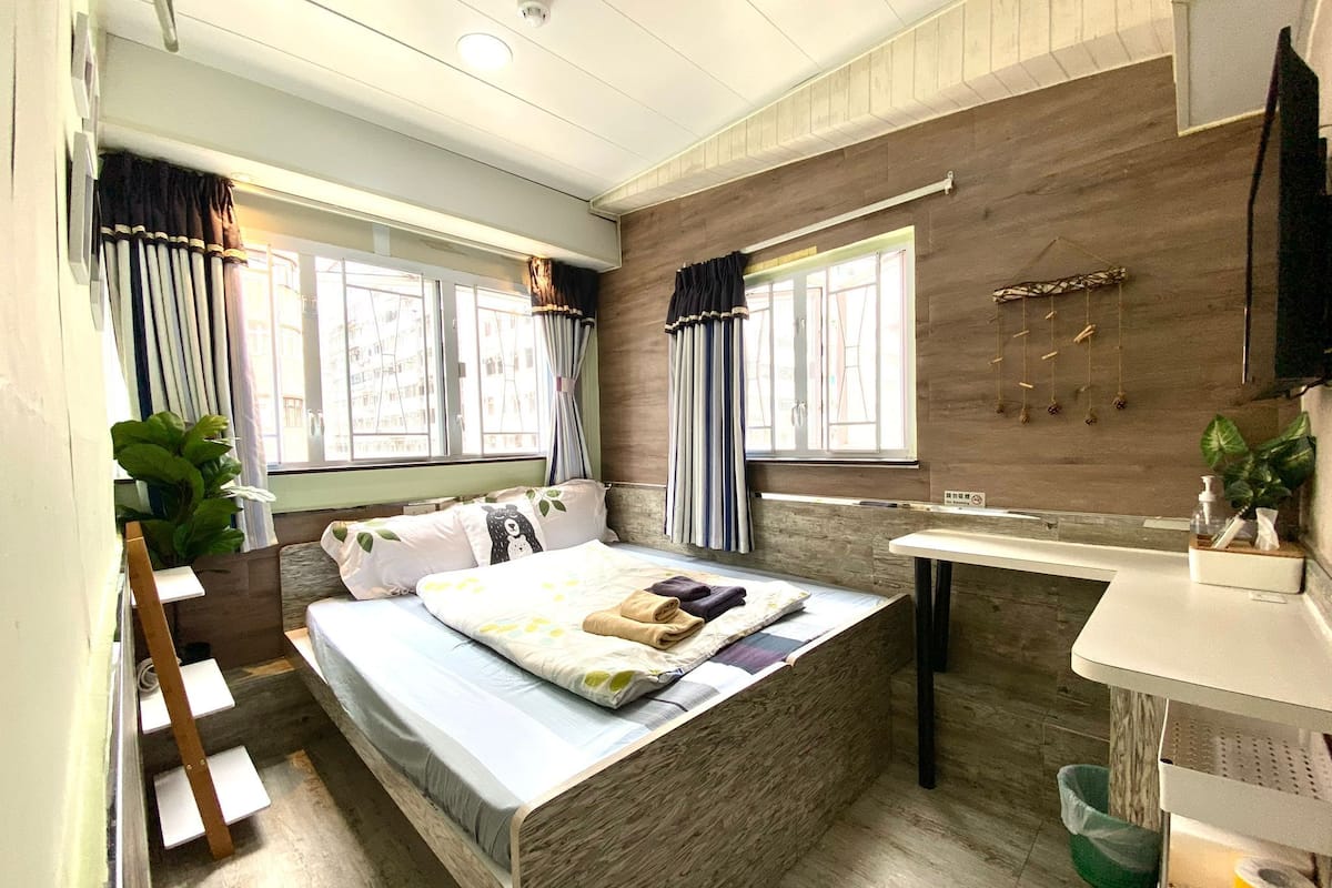 A bedroom with a bed, a bedside table, a window with curtains, a plant, and a wooden wall.
