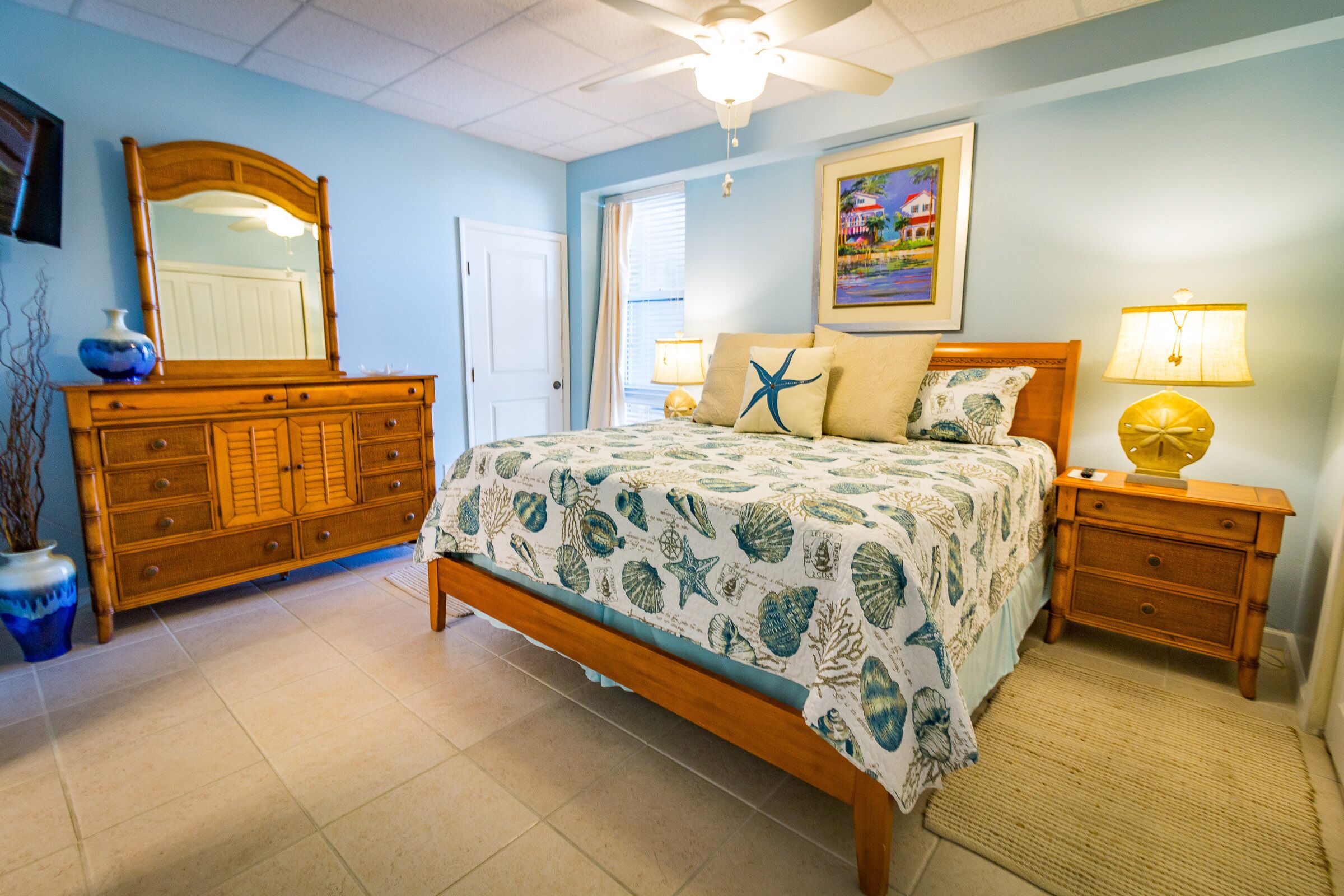 A bedroom with a bed, a dresser, a nightstand, a lamp, a mirror, and a painting on the wall.